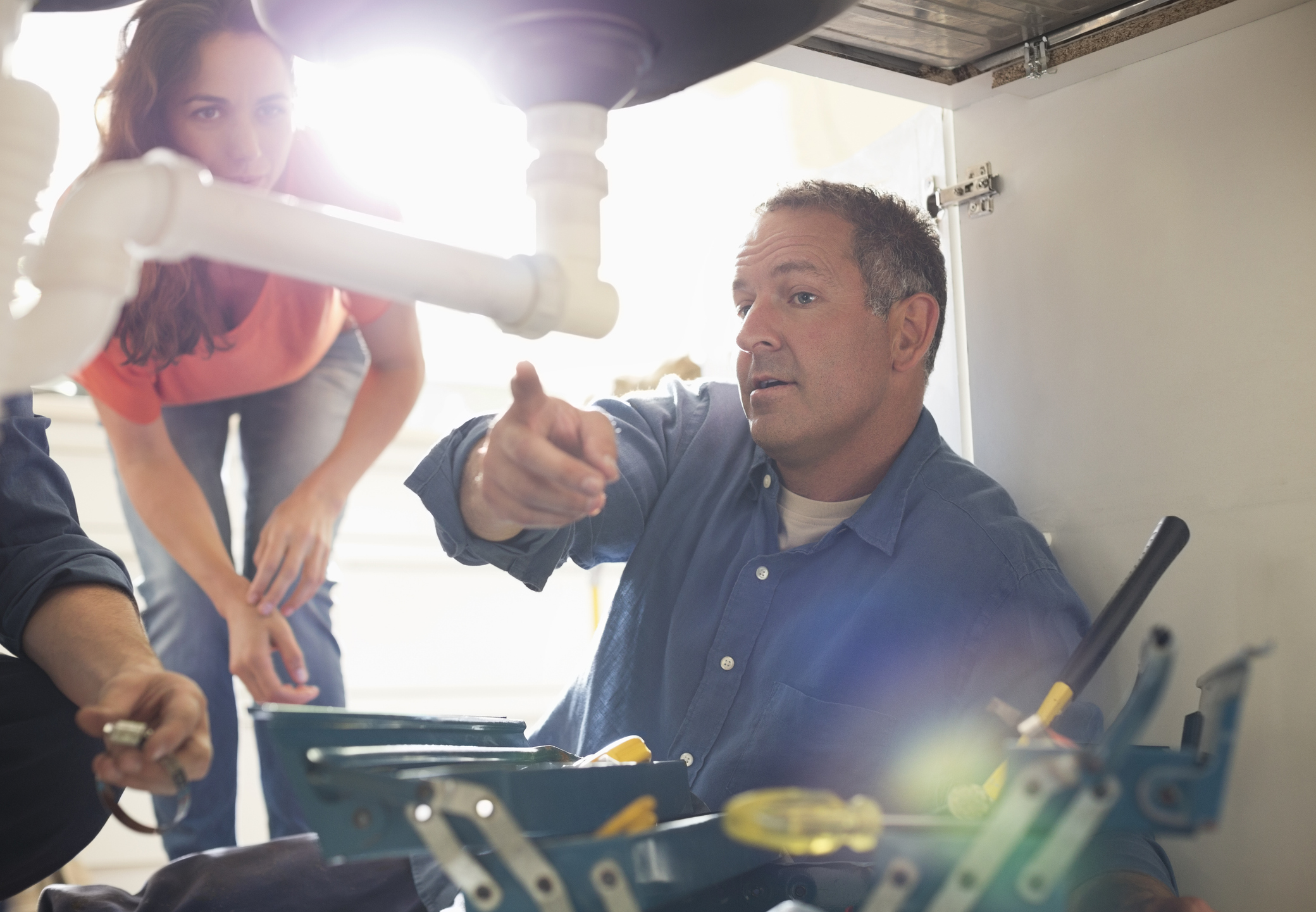 A man is fixing pipes under a sink while a woman watches. The man gestures toward the plumbing, explaining something. Tools are scattered nearby and sunlight streams in from behind them.