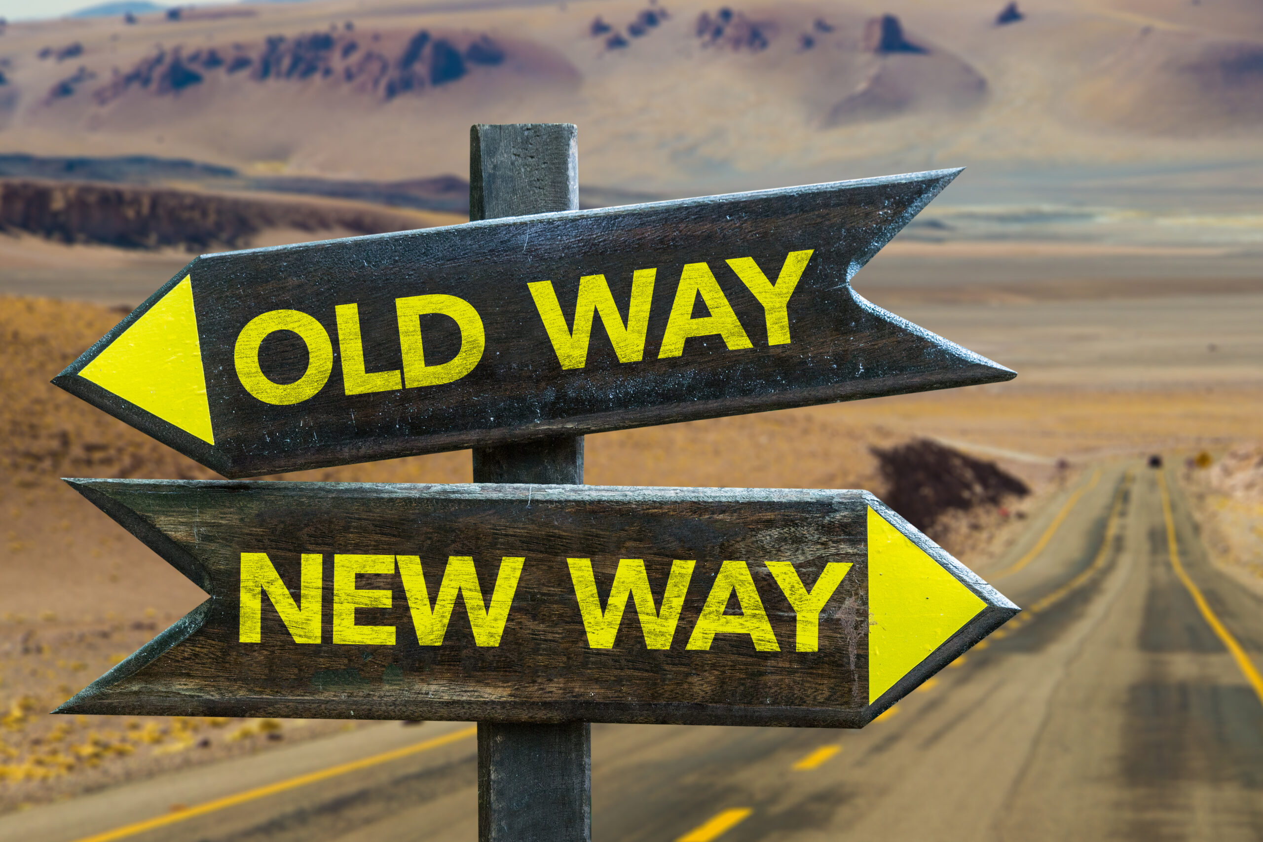Two wooden arrow signs at a crossroads in a desert, one pointing left labeled Old Way and the other pointing right labeled New Way, with a road stretching into the distant mountains.